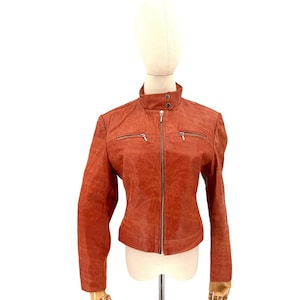 May include: A rust-colored leather jacket with a stand-up collar and a full-length zipper. The jacket has two zippered pockets on the chest and a tailored fit. The jacket is displayed on a mannequin.