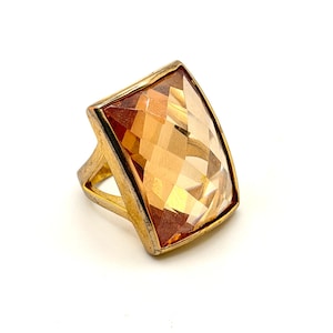 Könnte beinhalten: Ein goldfarbener Ring mit einem großen, rechteckigen, facettierten bernsteinfarbenen Stein. Die Fassung ist metallisch mit polierter Oberfläche und hält den Edelstein sicher in einem Rahmen, der den geometrischen Schliff und die warmen, orangefarbenen Töne betont.