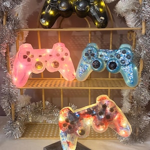 Pode incluir: Uma exibição de comandos de jogos em resina iluminados em designs preto, rosa, azul e multicolorido. Os comandos estão dispostos numa prateleira decorativa com detalhes em prata. Cada comando tem luzes embutidas.