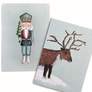 Op de afbeelding: Twee kerstkaarten met aquarel illustraties. Eén kaart toont een notenkraker soldaat in een teal uniform met een kerstboom. De andere kaart toont een rendier met verlicht gewei, in de sneeuw, op een lichtblauwe achtergrond.