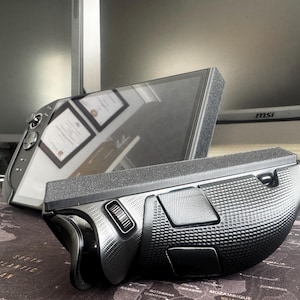 Könnte beinhalten: Eine schwarze und graue ergonomische Maus mit strukturierter Oberfläche und mehreren Tasten. Die Maus ist auf Komfort und Effizienz ausgelegt, mit einer geschwungenen Form und einem Scrollrad. Der Hintergrund umfasst einen Computermonitor und einen Schreibtisch.
