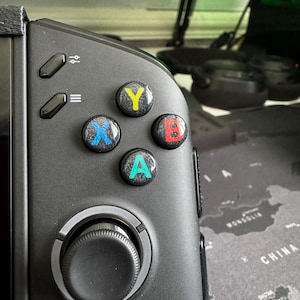 Puede incluir: Un mando de juego portátil negro con cuatro botones de acción circulares etiquetados como Y, X, B y A con una textura moteada. El botón Y es amarillo, X es azul, B es rojo y A es verde azulado. Dos botones de menú rectangulares están arriba.