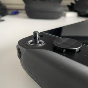 May include: Close-up of a black handheld gaming device. The device features a black joystick and a black, textured button. The device is on a white surface. The background is blurred.