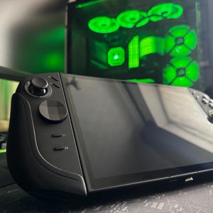 May include: A black handheld games console with a large screen and integrated controls. The device has a joystick, buttons, and a sleek, modern design. The background includes a computer keyboard and a desktop computer with green lighting.