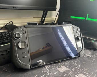 May include: A black handheld gaming console featuring a wide rectangular screen, integrated side controllers with analog sticks, a directional pad, and action buttons. The device is encased in a textured black frame with a top-mounted support bracket.