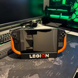 Könnte beinhalten: Ein schwarzes und orangefarbenes Handheld-Gaming-Gerät mit dem Wort "LEGION" in weißen Buchstaben und einem roten Kreislogo. Das Gerät steht auf einem schwarzen Ständer, der auf einem Weltkarten-Mauspad ruht.