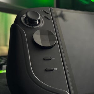 May include: Close-up of a black handheld gaming device. The device features a joystick, a circular directional pad, and several buttons. The screen is black and appears to be turned off. The device has a sleek, modern design.