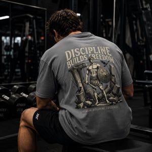 Könnte beinhalten: Ein graues Kurzarm-T-Shirt mit einem Rückendruck, der den Text "DISCIPLINE BUILDS FREEDOM" und "STRENGTH. DISCIPLINE. LIFESTYLE." zeigt. Die Illustration stellt einen Krieger mit Schild und Schwert zwischen Steinsäulen dar.