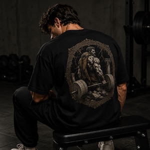 Może przedstawiać: Czarny t-shirt z krótkim rękawem z dużą, okrągłą grafiką na plecach. Wzór przedstawia muskularną postać w skórze lwa, podnoszącą ciężką sztangę wewnątrz ozdobnej ramy z greckimi wzorami i kamiennymi kolumnami w tle.