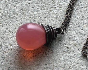 May include: A necklace featuring a smooth, translucent pink spherical bead pendant. The bead is secured at the top with dark, oxidized metal wire wrapping that connects to a matching dark metal chain with small, uniform circular links.