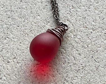 May include: A teardrop-shaped red glass pendant suspended from a dark metal chain. The top of the smooth, matte-finish red glass bead is secured with a decorative wrapping of thin dark metal wire that connects directly to the link chain.
