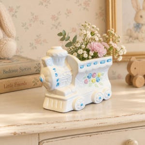 May include: A white ceramic train-shaped planter featuring blue accents on the wheels and top, decorated with a small cluster of pastel-colored sculpted roses on the side, currently holding a bouquet of white baby's breath and pink carnations.