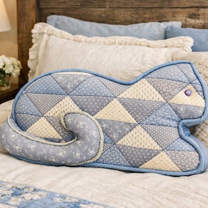 May include: A cat-shaped cushion with a patchwork design in shades of blue and cream. The cushion features a blue border and a curled tail. The patchwork is made of triangular fabric pieces. A purple bead is used for the eye. The cushion is on a bed.