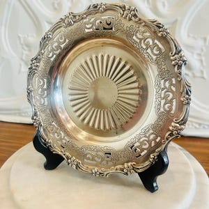 Peut inclure: Une assiette décorative argentée avec un motif floral et des volutes ornementales sur le bord. L'assiette présente un motif central en forme d'éventail et est présentée sur un support noir, sur fond blanc.