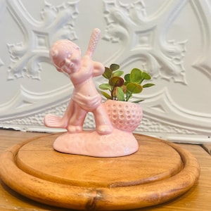May include: A pink ceramic figurine of a baby golfer with a golf ball planter holding small green plants. The figurine is on a wooden round tray.