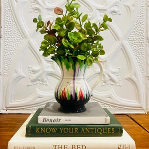 May include: A decorative, hand-painted ceramic pitcher with a black base, white body, and colorful accents, holding green and red-tinged faux foliage. The pitcher sits atop a stack of three books: "Renoir", "Know Your Antiques", and "The Bed".