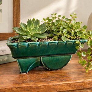 May include: A glossy, dark green ceramic planter shaped like a miniature wheelbarrow. The planter holds a green succulent and a trailing green plant with small, rounded leaves. The surface of the ceramic has a smooth, reflective finish.