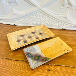 May include: Two rectangular ceramic dishes with a speckled, tan base. One has a checkered pattern, the other floral designs in blue, green, and pink. Both are on a wooden surface. The dishes are likely for serving.