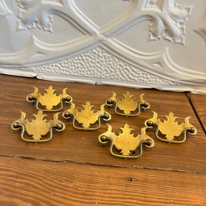 May include: A set of five gold-toned metal drawer pulls, each featuring an ornate, symmetrical backplate with a central decorative finial and a curved, hinged drop handle attached to two small circular mounting points on the sides.