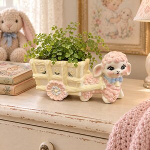 May include: A ceramic planter shaped like a pink lamb pulling a pale yellow cart with a woven texture and small flower details. The lamb has large dark eyes, a blue bow around its neck, and a textured coat. A green fern plant sits inside the cart.
