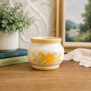 May include: A cream-coloured ceramic planter with a rounded body and a flared rim. The exterior features a painted floral pattern of bright yellow and light blue flowers with green leaves, accented by horizontal yellow bands at the top and base.