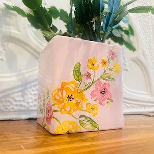 May include: A pale pink ceramic vase featuring a floral design in yellow, pink, and red. The flowers are painted in a watercolour style, with green leaves. The vase is square-shaped and sits on a wooden surface.