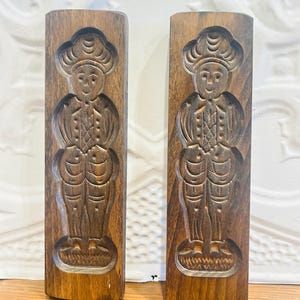 May include: Two vintage wooden cookie molds. Each mold is rectangular and features a carved image of a person in traditional attire. The wood has a warm, brown tone, and the carvings are detailed, showing the figure's clothing and facial features.
