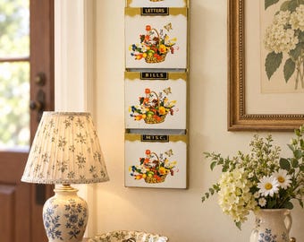 May include: A vertical wall-mounted metal organizer featuring three stacked pockets labeled LETTERS, BILLS, and MISC. Each pocket is decorated with a colorful illustration of a woven basket filled with assorted flowers and fruits.