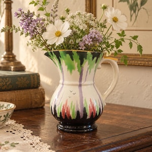 May include: A ceramic pitcher featuring a white base with a painted pattern of green, purple, and red vertical brushstrokes, a black base, and a small handle. The pitcher is filled with a bouquet of white cosmos, purple flowers, and green foliage.