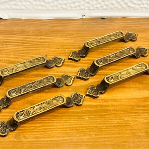 May include: A set of five metal drawer pulls featuring a dark finish with gold-toned, embossed floral patterns on the top surface. Each handle has a curved shape with decorative, flared ends and two mounting points for attachment to furniture.