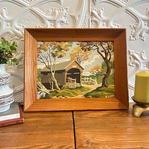 May include: A framed paint-by-number landscape painting featuring a covered bridge, trees with autumn-toned foliage, and a stone wall. The artwork is enclosed in a medium-toned wooden frame and displays the signature R. SYREK in the bottom corner.