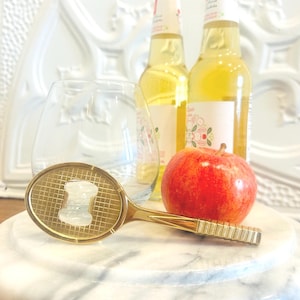 May include: A gold-coloured bottle opener shaped like a tennis racket, a red apple, two bottles of sparkling cider, and a wine glass are arranged on a marble serving board. The bottles have white labels with floral designs.