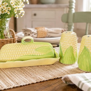 Peut inclure: Un beurrier en céramique et une paire de salière et poivrière, tous conçus pour ressembler à des épis de maïs avec des grains jaunes et des feuilles vertes. Le beurrier possède un couvercle amovible avec une anse, et les shakers sont cylindriques.