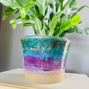 Peut inclure: Un pot de fleurs en céramique avec un vernis brillant présentant des bandes horizontales turquoise, bleues et violettes, se terminant par une base beige brute non émaillée. Une plante Schlumbergera aux feuilles plates pousse dans le pot.