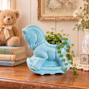 May include: A light blue ceramic rocking horse planter with a glossy finish. The hollow back of the horse holds a small green plant with trailing stems and round leaves. The horse is shaped in a classic rocking design with a curved base.