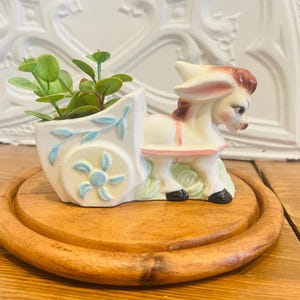 Peut inclure: Une figurine d'âne en céramique tirant une charrette jardinière, peinte en crème avec des motifs floraux bleu clair et des détails de harnais roses. L'âne a des cheveux bruns et des sabots noirs, et la charrette contient une petite plante verte.