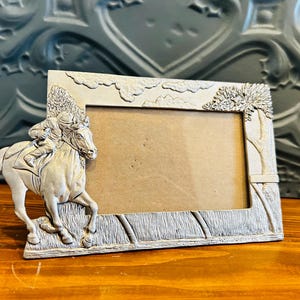 May include: A rectangular, off-white picture frame with a detailed relief of a horse and jockey on the left side. The frame has a textured border with cloud and tree details. The frame is made of a light-colored metal.