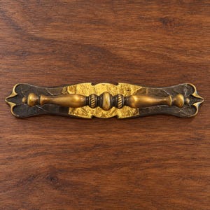 May include: A horizontal drawer pull featuring a central brass-toned handle with a rope-textured middle section. The handle is mounted on a decorative backplate with a dark, textured finish and a central panel of bright, metallic gold leaf.