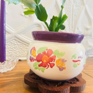 May include: A small, round, white ceramic planter with a deep purple rim, decorated with a watercolour-style floral pattern in red, orange, and green. The planter sits on a dark brown, carved wooden coaster, holding a green succulent plant.