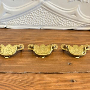 Peut inclure: Trois poignées de tiroir en laiton composées d'une plaque arrière semi-circulaire avec un motif floral gravé et d'une poignée pivotante assortie. Chaque pièce présente une finition dorée métallique et un design incurvé avec des détails gravés.
