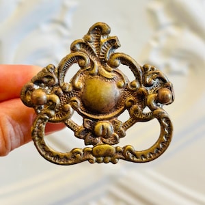 May include: A brass-toned metal drawer pull featuring an ornate, symmetrical design with a central circular medallion, intricate scrollwork, and a hinged drop handle. The finish has a weathered, antique appearance with darker patina accents.