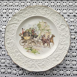 May include: A decorative white plate with a gold rim and embossed floral patterns. The center features a detailed illustration of a horse-drawn carriage with passengers. The plate's design evokes a vintage aesthetic, suitable for display or dining.