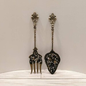 May include: A set of two ornate silver-toned serving utensils with intricate designs. The fork and spoon have decorative handles with a fleur-de-lis design.