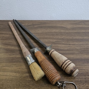 May include: Three kitchen tools on a wooden surface. A small brush with a wooden handle and metal ferrule, a knife sharpener with a wooden handle, and a steel rod with a wooden handle and metal guard.