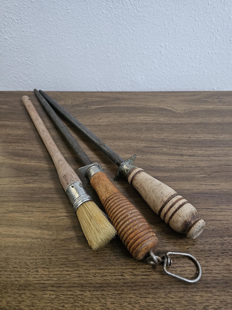 May include: Three kitchen tools on a wooden surface. A small brush with a wooden handle and metal ferrule, a knife sharpener with a wooden handle, and a steel rod with a wooden handle and metal guard.