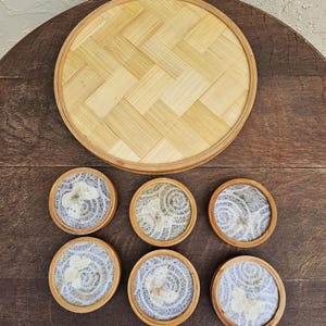 K&ouml;nnte beinhalten: Ein Set aus sechs runden Untersetzern mit hellblauem und wei&szlig;em Design, jeweils in einem Rahmen aus Naturholz. Ein passendes rundes Bambustablett befindet sich &uuml;ber den Untersetzern. Die Untersetzer und das Tablett liegen auf einer dunklen Holzoberfl&auml;che.