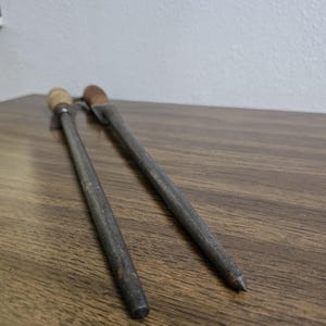 May include: Two metal knife sharpeners with wooden and brown handles rest on a wooden surface. The sharpeners are gray and have a cylindrical shape, with one ending in a point and the other with a flat end.