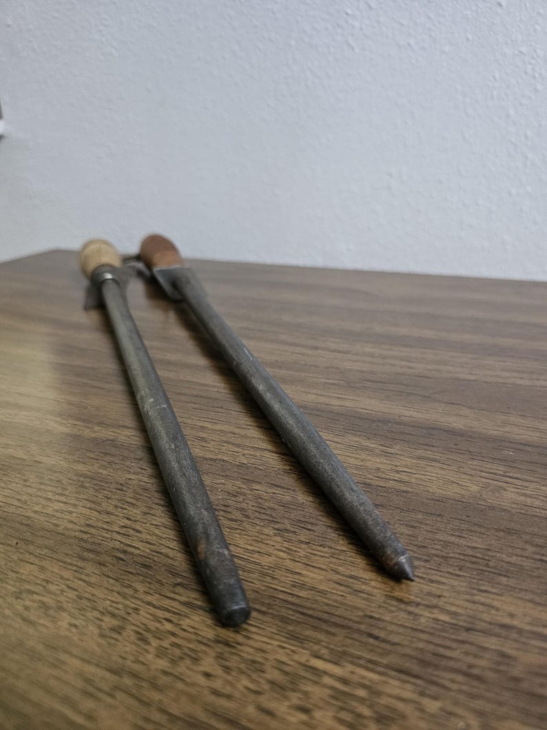 May include: Two metal knife sharpeners with wooden and brown handles rest on a wooden surface. The sharpeners are gray and have a cylindrical shape, with one ending in a point and the other with a flat end.