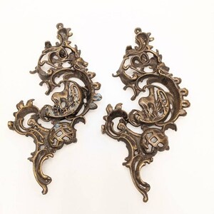 May include: Two ornate, antique bronze-colored metal decorative furniture appliques. Each features a scrolling, symmetrical design with floral and foliate elements, and a small animal figure. The appliques are set against a white background.
