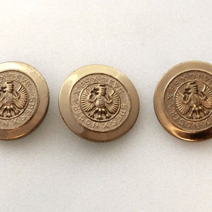 May include: Three antique gold-colored buttons with an embossed eagle design and Latin text. The buttons are round and have a slightly aged appearance, suggesting they are vintage or vintage-inspired.